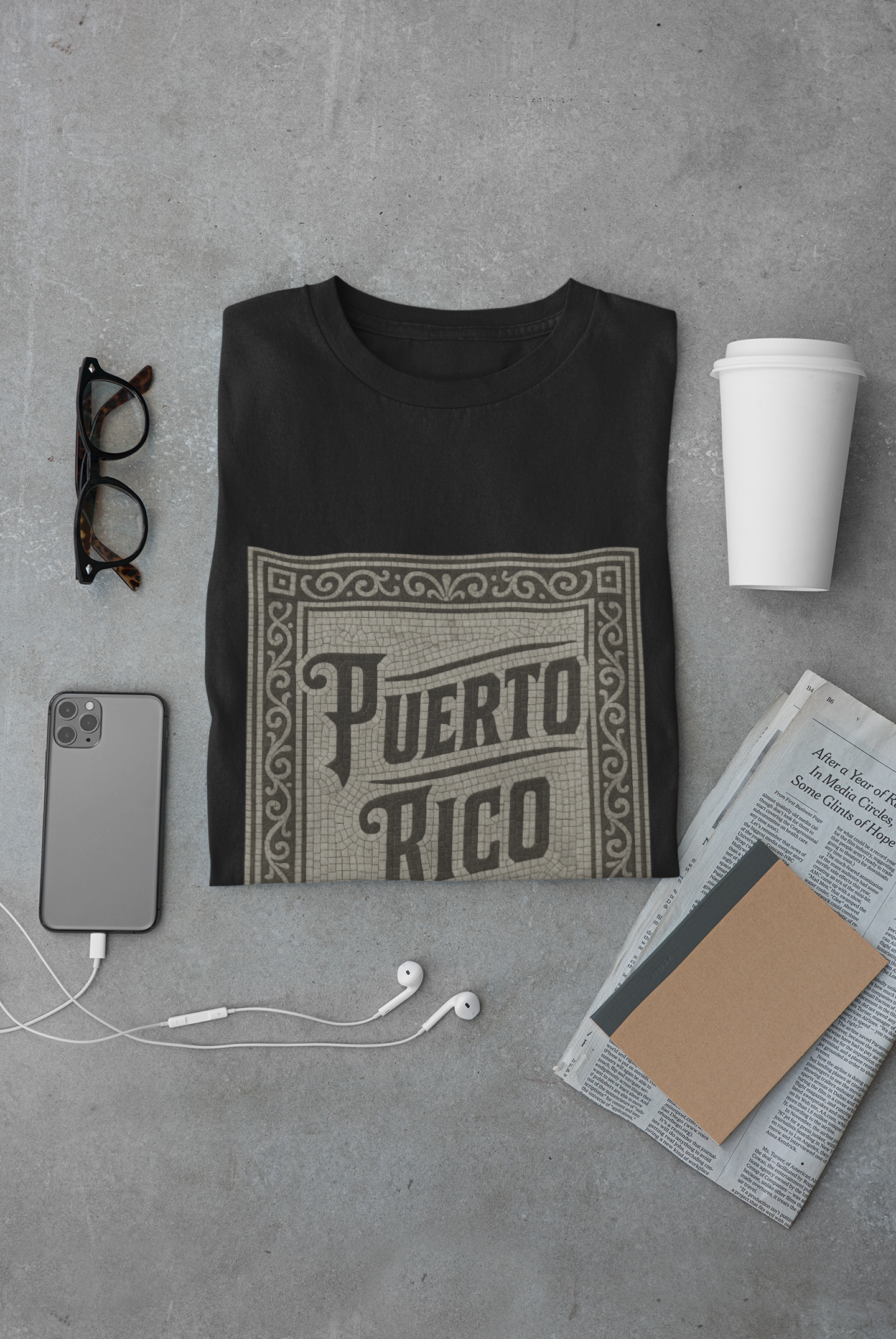 Black t-shirt with 'Puerto Rico' design on a gray surface with phone, glasses, and newspaper.